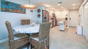 Un comedor con una mesa redonda y sillas de mimbre, una cocina al fondo y un cuadro con motivos playeros en la pared.