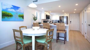 A dining area with a round table and four chairs, a kitchen in the background, and a door leading to another room.