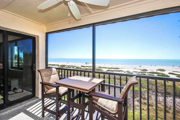 A balcony with a table and chairs, overlooking a beach with the ocean and a clear sky.