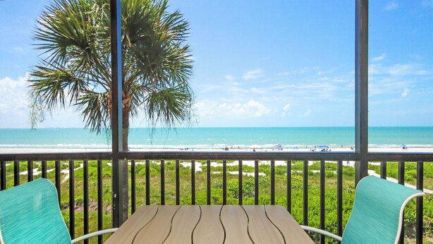 Ein Holztisch mit zwei Stühlen auf einem Balkon mit Blick auf einen Strand, Palmen und einen klaren Himmel.