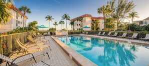 A swimming pool with lounge chairs and umbrellas, surrounded by palm trees and a building with a red roof.