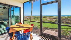 Un balcón con una mesa de madera y sillas coloridas, con vista a una playa y palmeras.