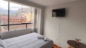 A hotel room with a bed, a small table, a TV mounted on the wall, and a window with a view of buildings and mountains. - Hostal El Mariscal  (Urubamba)