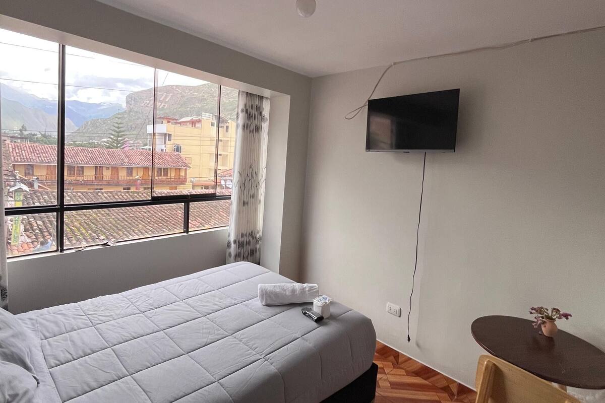 A hotel room with a bed, a small table, a TV mounted on the wall, and a window with a view of buildings and mountains.
