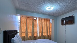 A bedroom with a bed, curtains, a ceiling light, and a TV mounted on the wall. - Hostal El Mariscal  (Urubamba)