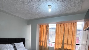 A neatly made bed with a grey bedspread and white pillows, a window with orange curtains, and a ceiling light. - Hostal El Mariscal  (Urubamba)