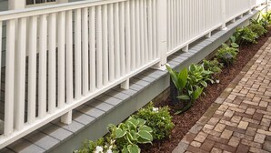 Une véranda en bois peinte en blanc, avec un escalier menant à une porte, le tout entouré d’un chemin en briques et de petites plantes.