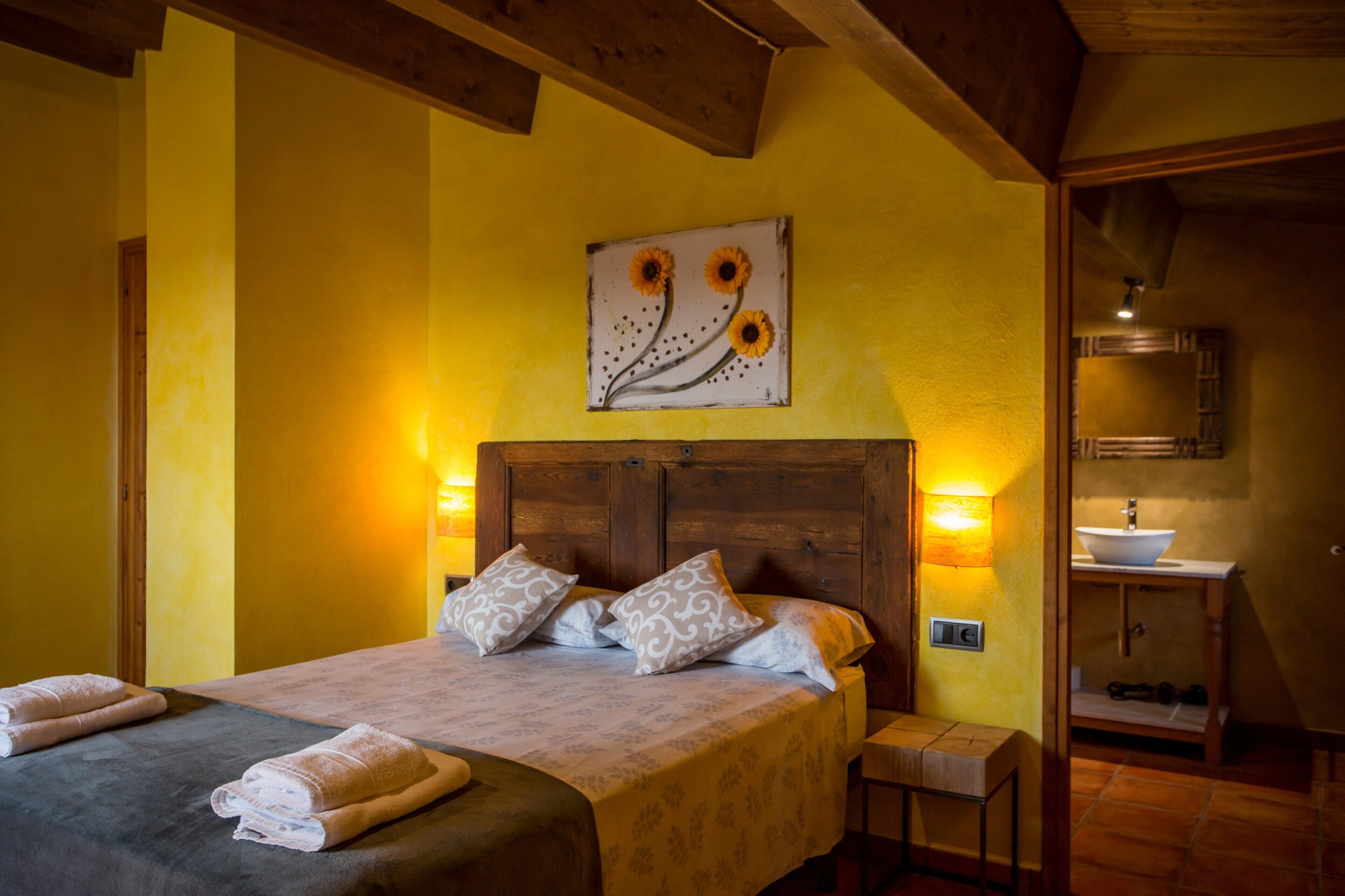 Un dormitorio con cama, almohadas, una mesita de noche, una obra de arte en la pared y un baño con lavamanos y espejo.