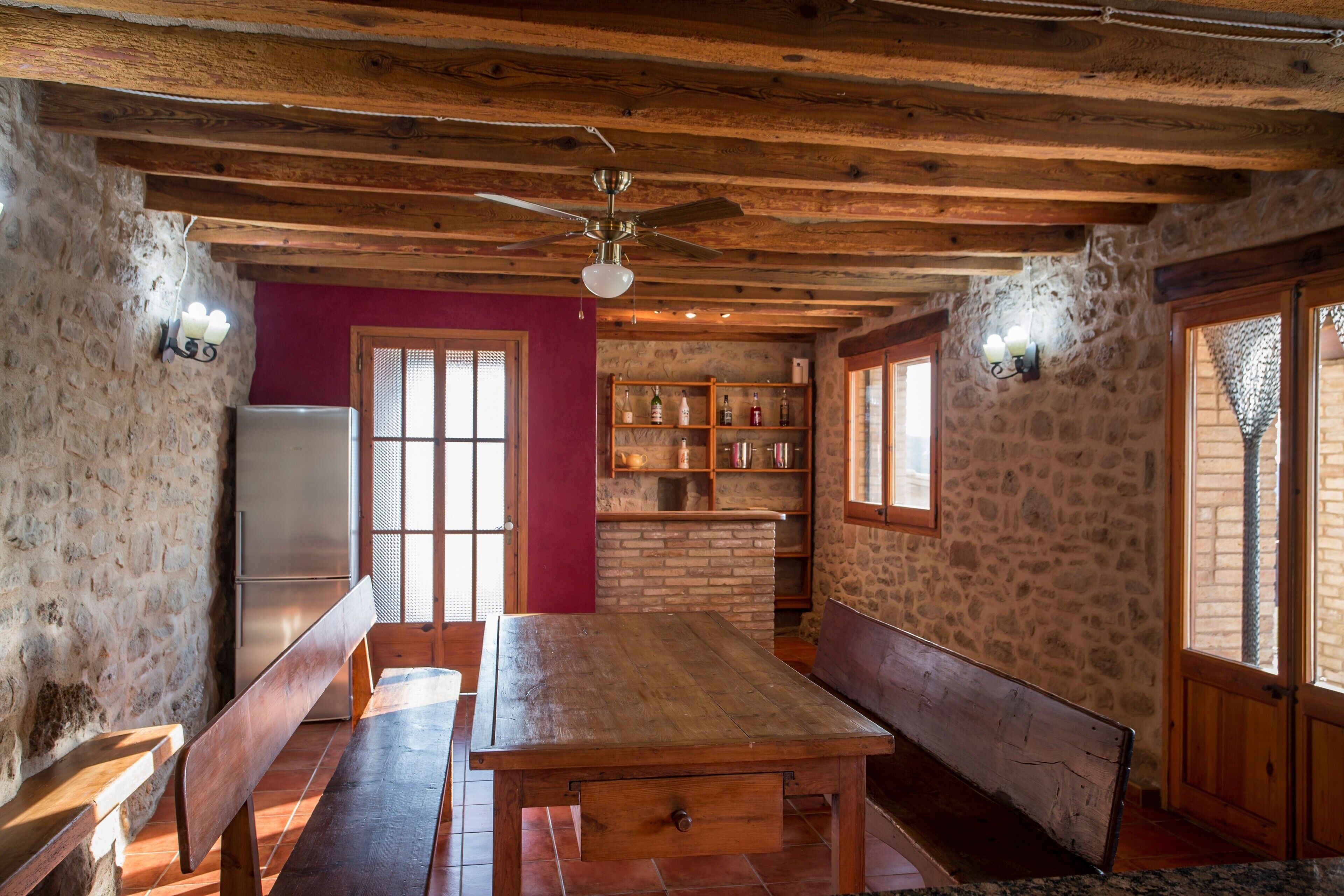 Un comedor rústico con una mesa y bancos de madera, ventilador de techo, paredes de piedra y chimenea de ladrillo.