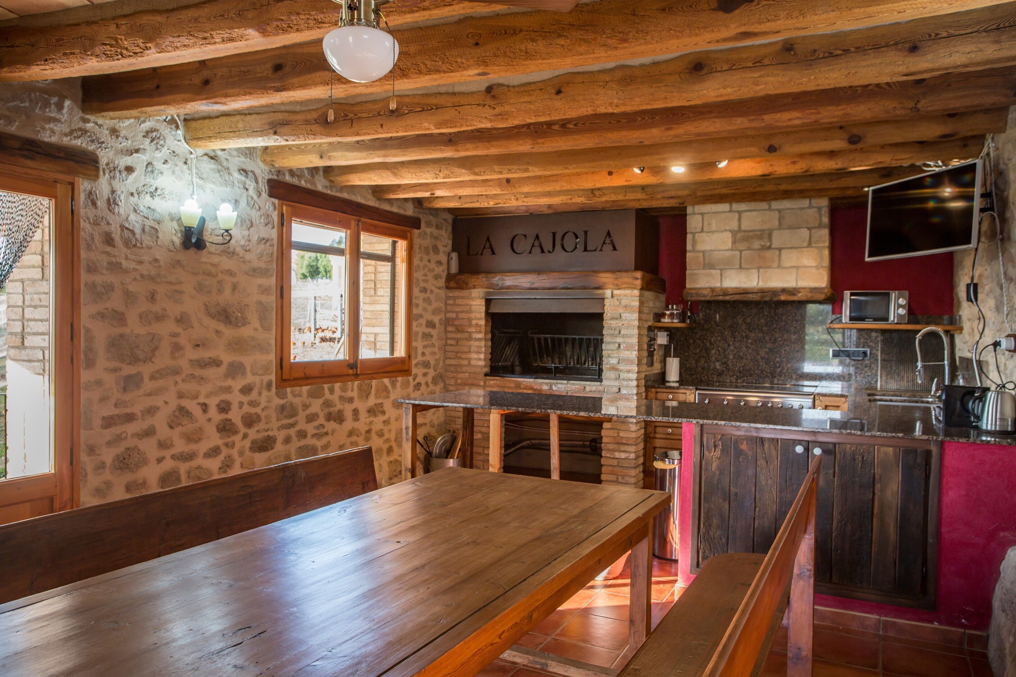 Cocina rústica con pared de piedra, vigas de madera, una mesa de madera y un cartel que dice 'LA CAJOLA'.