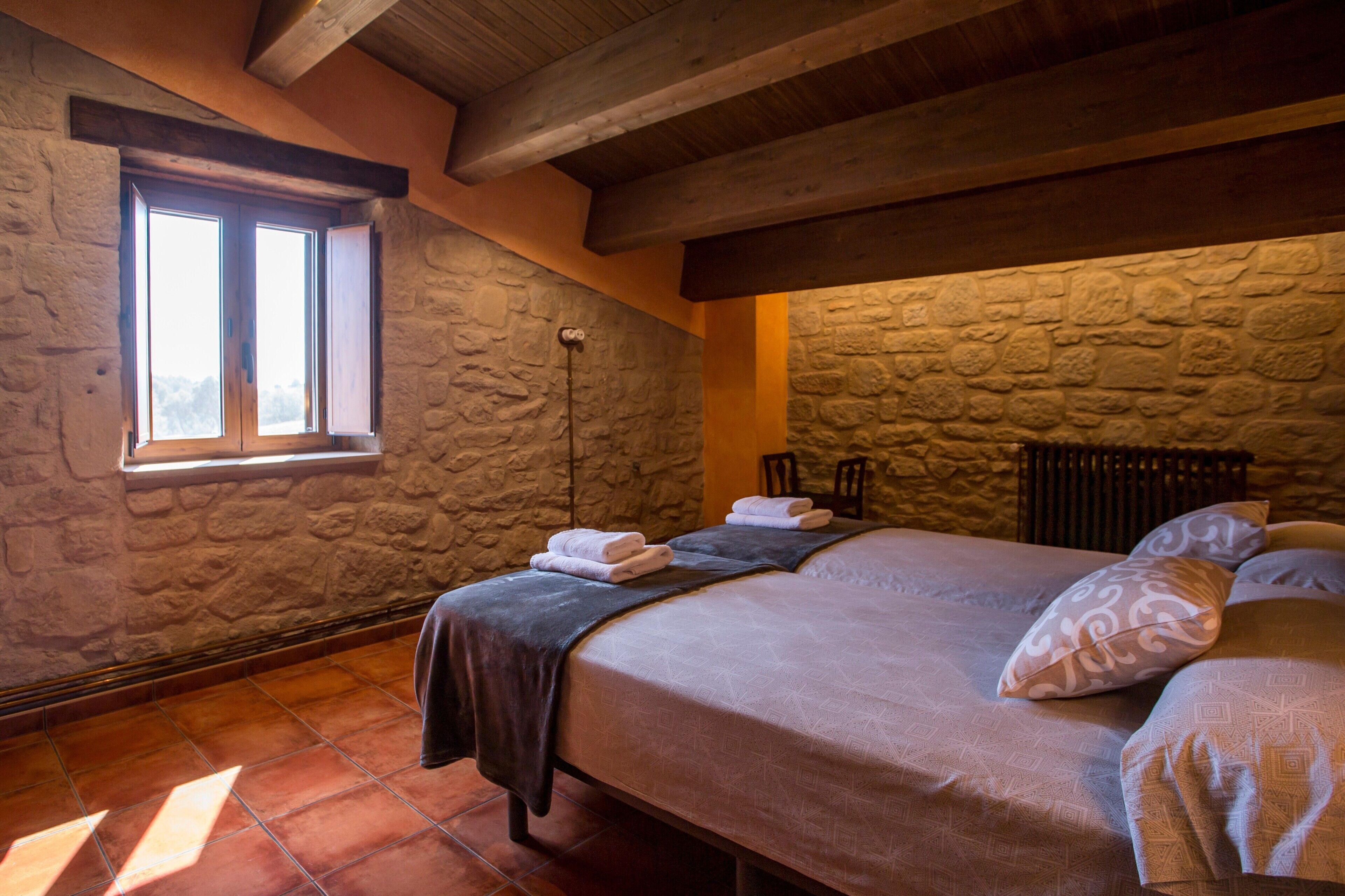 Een kamer met een stenen muur, een bed met een donkere dekbedovertrek, een raam met open luiken en een stoel.