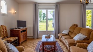 Una sala de estar con televisor, sofá y vista a un jardín a través de grandes ventanales.