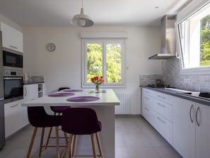 Una cocina moderna con armarios blancos, una isla central con individuales morados, una estufa a gas y una ventana con vistas al verdor.