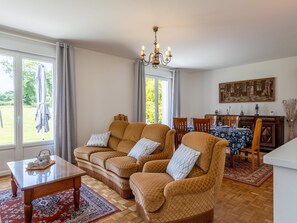 Una sala de estar con dos sillones, una mesa de centro, una araña, una mesa de comedor con sillas y un cuadro en la pared.