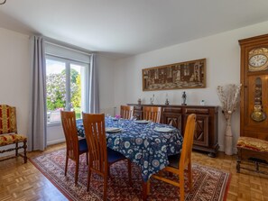 Un comedor con una mesa preparada para cuatro, sillas, un mantel estampado, un cuadro grande, un reloj y vistas a la vegetación exterior.