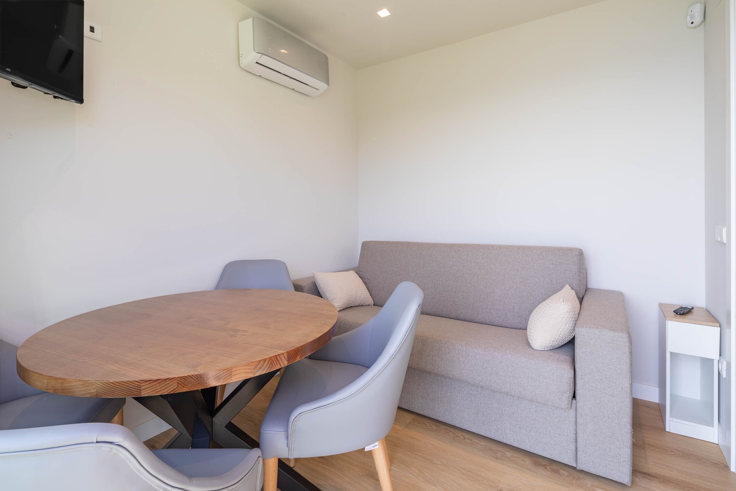 A room with a round wooden table, two grey chairs, a grey sofa with cushions, a white side table, and a mounted TV.