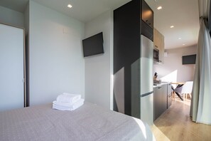 A hotel room with a bed, a flat-screen TV mounted on the wall, a kitchenette, and a dining area.
