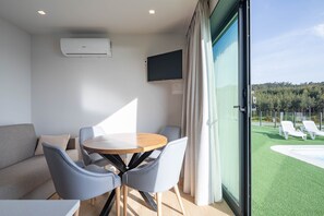 A modern room with a round table, chairs, a sofa, a TV, and a large window with a view of a pool and greenery.