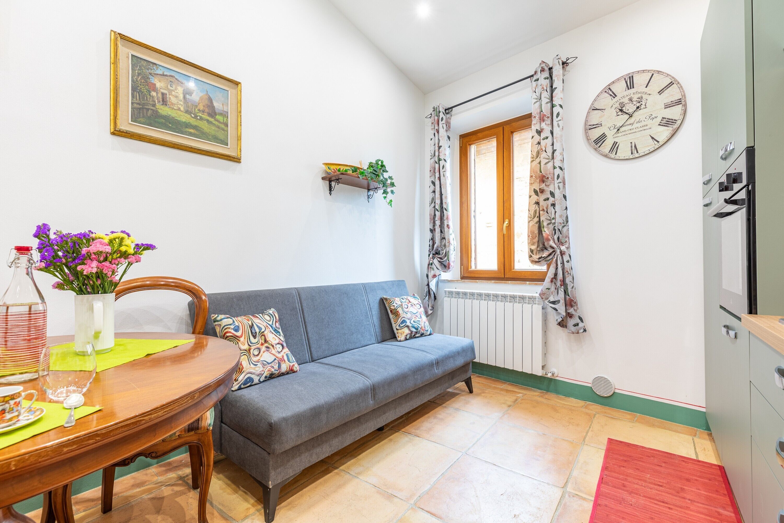 A living room with a sofa, a dining table with a vase of flowers, a clock, and a window with curtains.