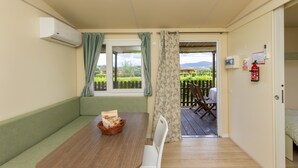 Ein Zimmer mit einem Holztisch, einer gepolsterten grünen Sitzbank, einem Korbwäscher, einem Fenster mit Vorhängen, einer Tür zu einem Außenbereich mit einer Holzterrasse und einem an der Wand befestigten Feuerlöscher.