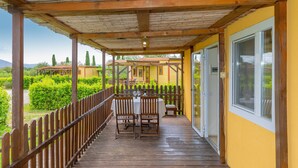 Eine überdachte Terrasse im Freien mit Holztisch und -stühlen, einem Holzgeländer und Blick auf ein gelbes Gebäude.