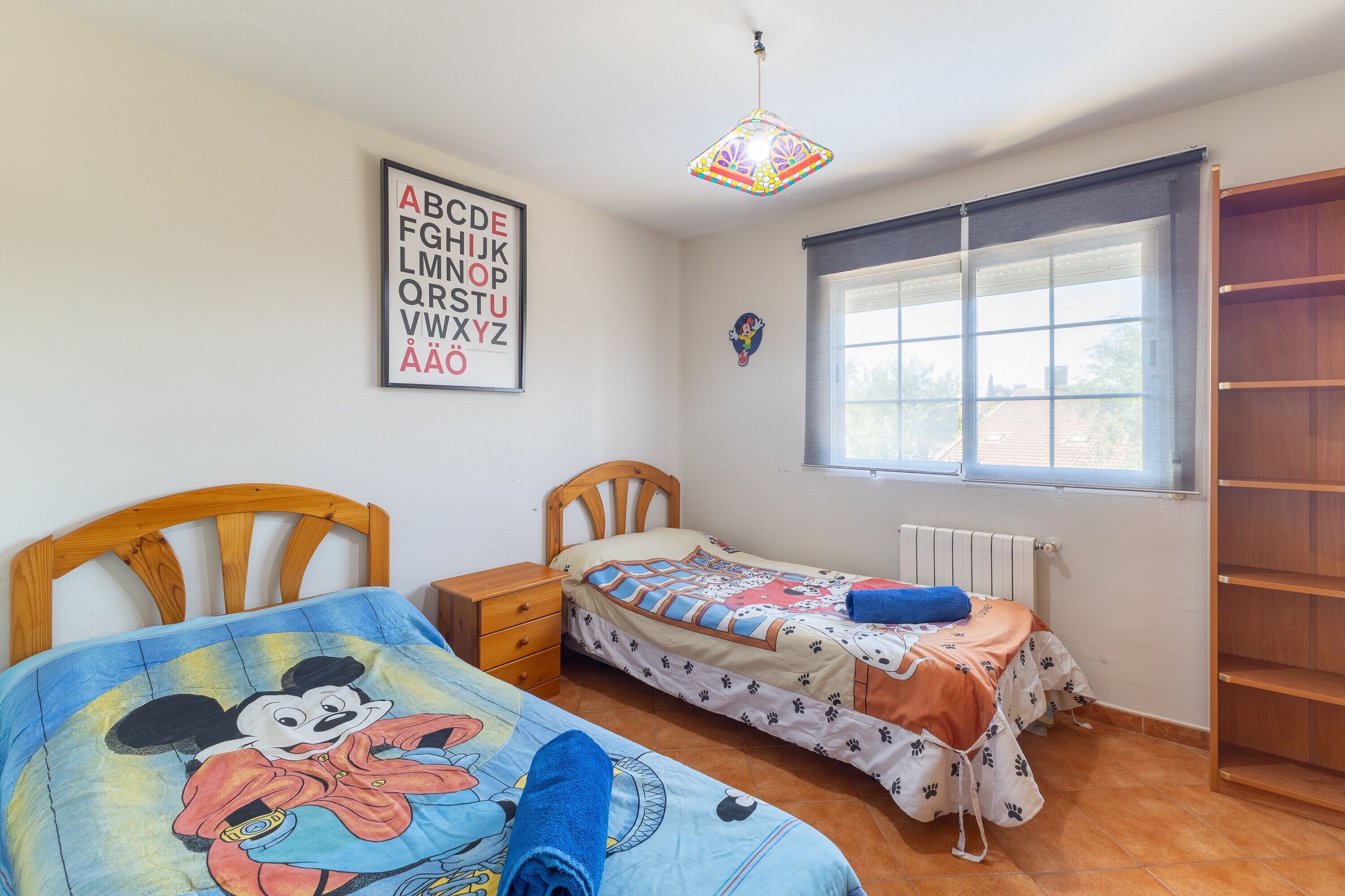 Un dormitorio con dos camas individuales, una ventana con persianas, un radiador, un armario de madera y un cartel del alfabeto colgado en la pared.