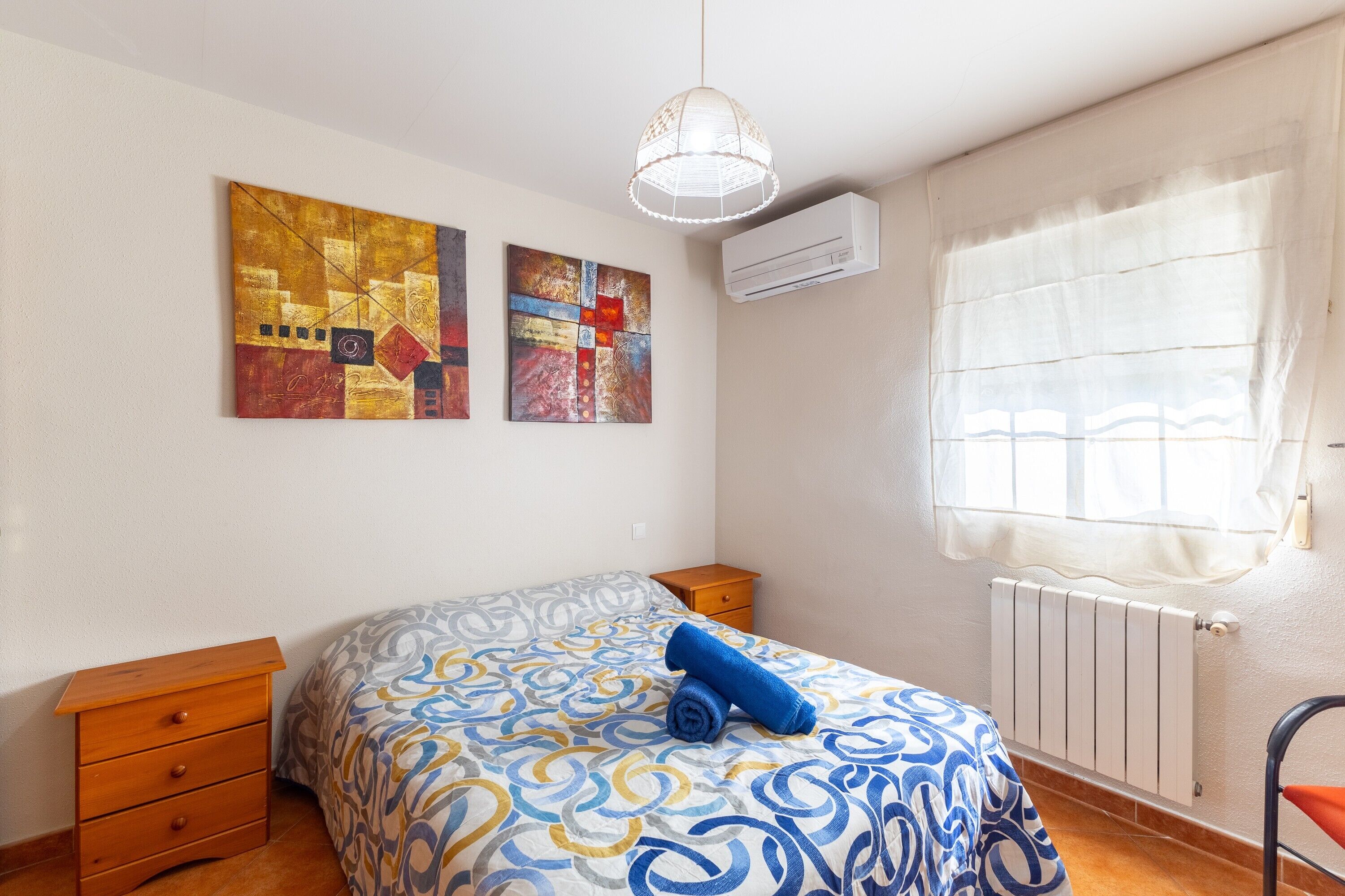A bedroom with a bed, two paintings, a radiator, a window with curtains, and a wooden nightstand.