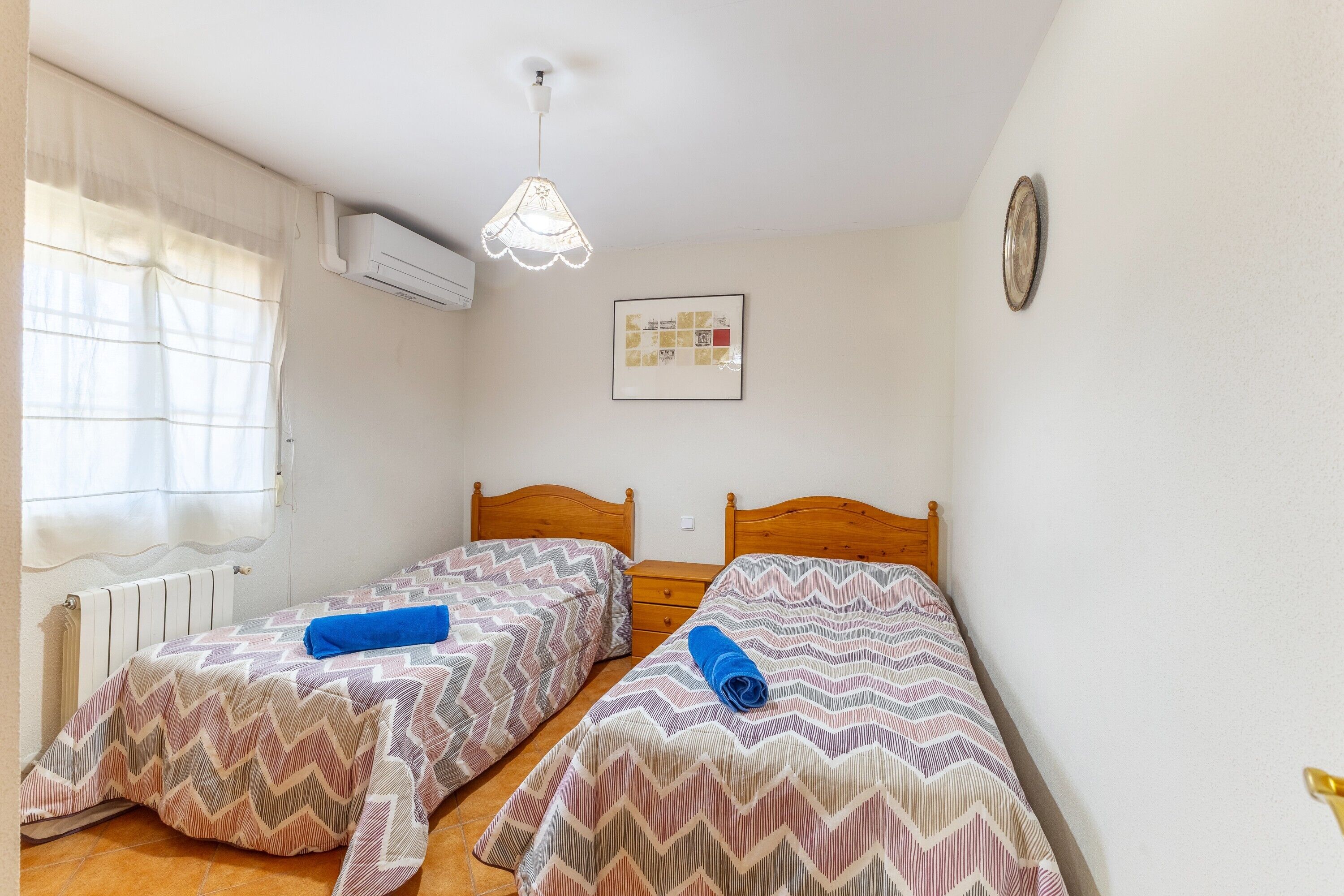 Quarto pequeno e limpo com duas camas, janela com cortinas, ar condicionado, relógio de parede e um quadro emoldurado.