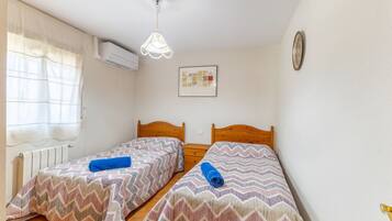 Quarto pequeno e limpo com duas camas, janela com cortinas, ar condicionado, relógio de parede e um quadro emoldurado.
