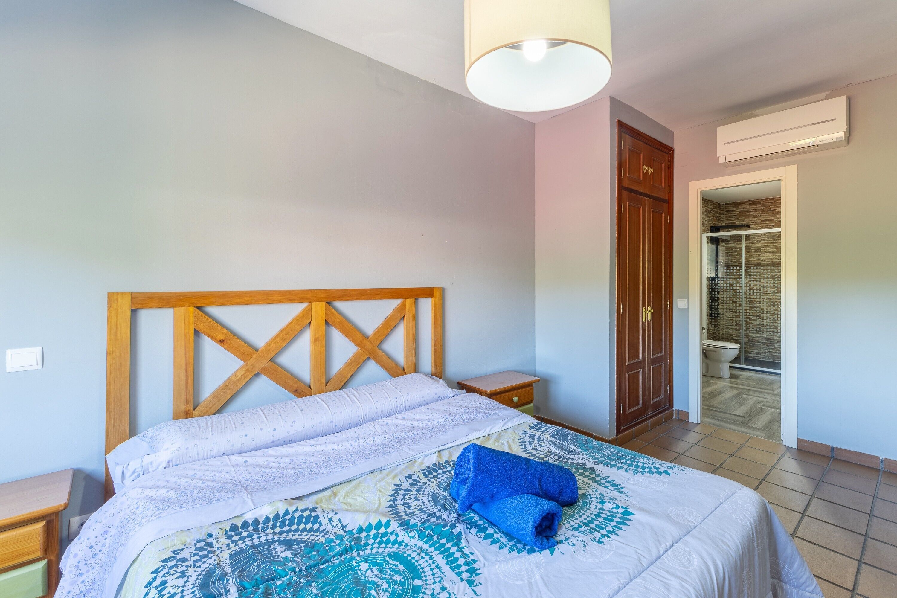 Un dormitorio con cabecera de madera, cama con ropa de cama estampada, mesita de noche, armario y aire acondicionado.