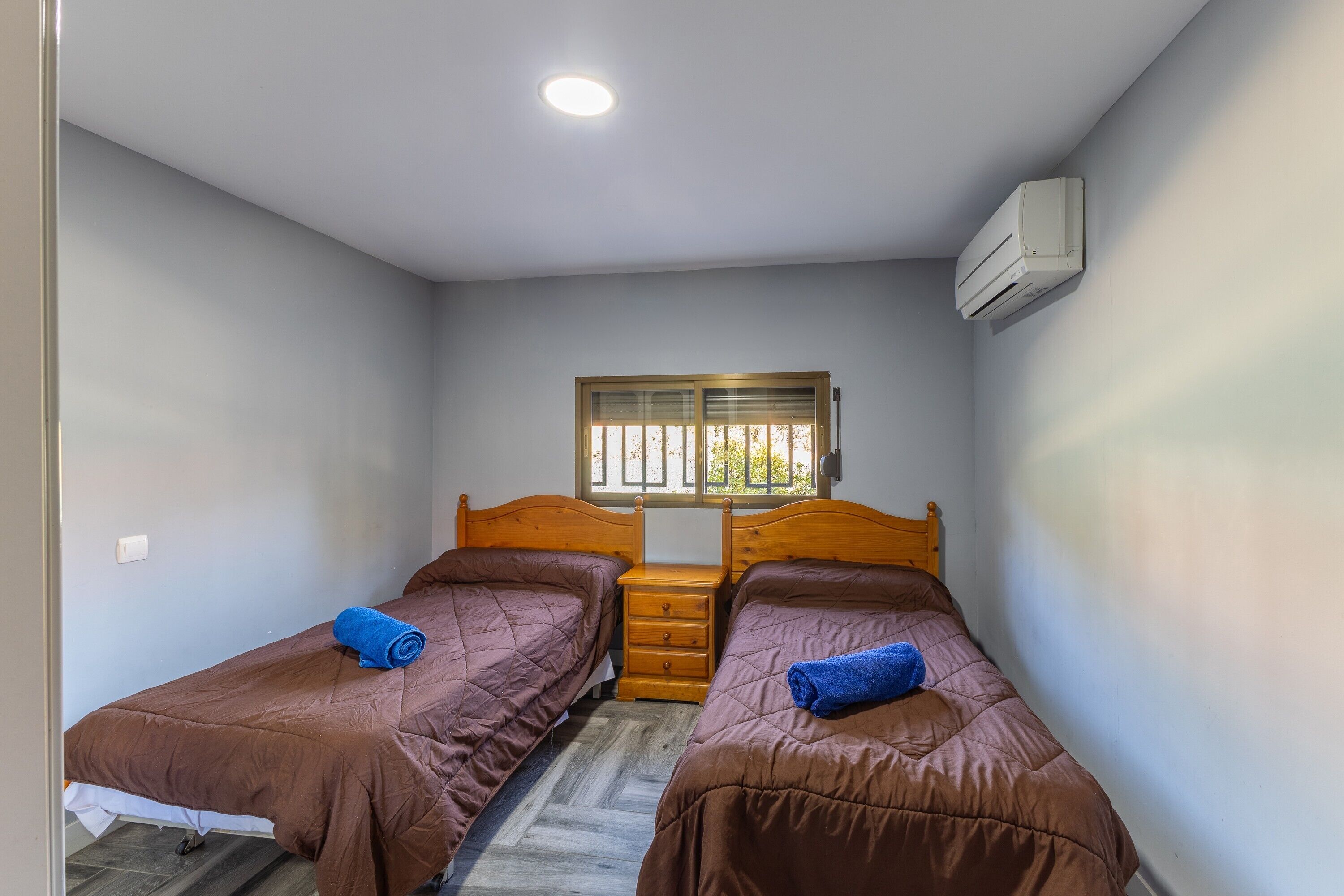 A small, single-room accommodation with two beds, a window, a wooden nightstand, and an air conditioning unit.