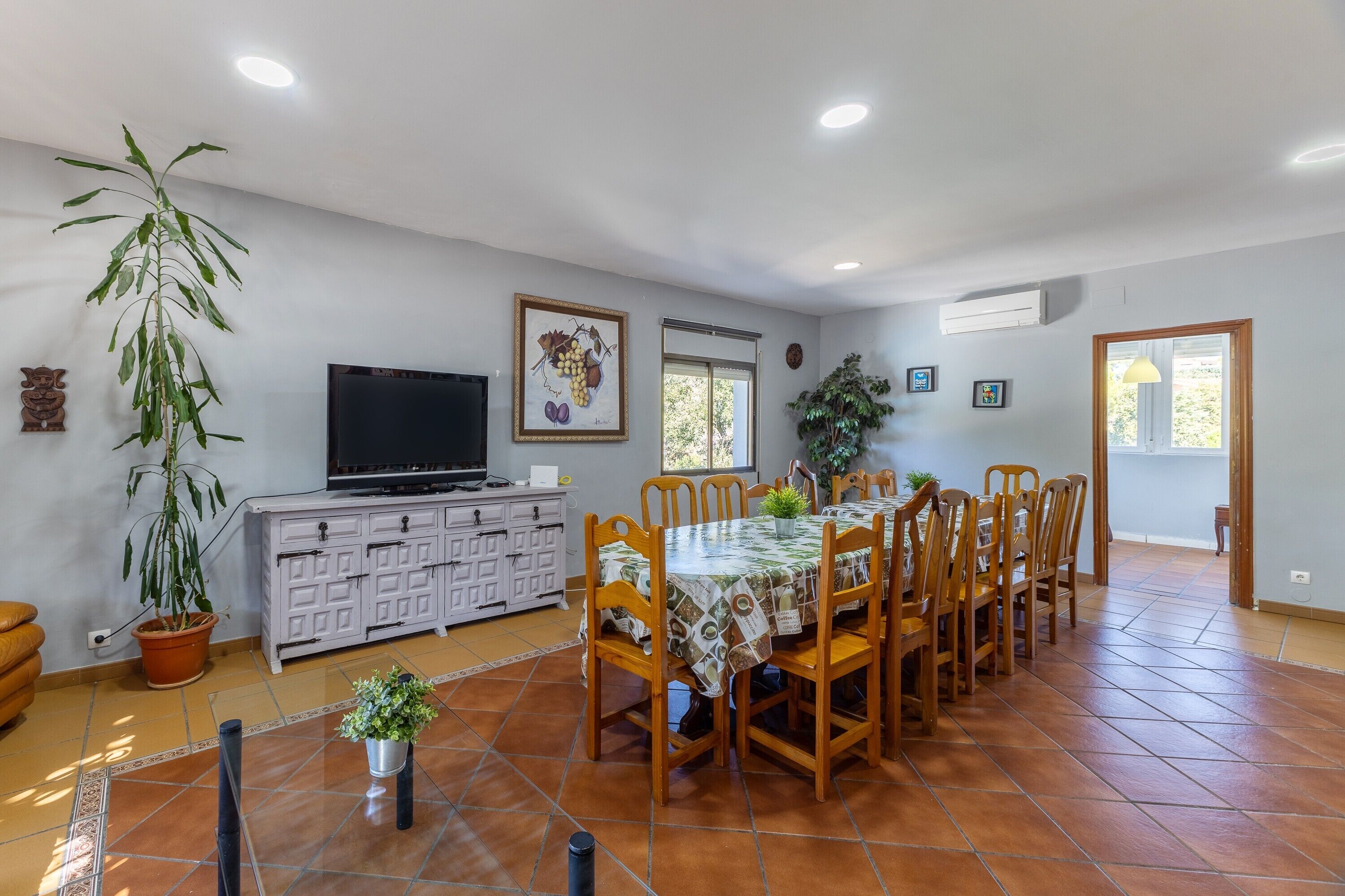 A dining area with a table set for six, surrounded by chairs, a television on a cabinet, and potted plants.