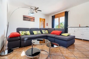 Un salon comprenant un canapé d’angle, un ventilateur de plafond, une petite table ronde et une vue sur l’extérieur par une fenêtre.