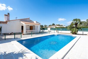Une piscine avec une terrasse carrelée de couleur blanche, un petit bâtiment doté d’un toit en tuiles et un palmier en arrière-plan.
