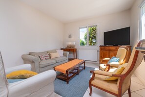 A living room with a sofa, armchair, coffee table, TV, and a window with a view of trees. - Serene Getaway Steps from the Carnac Alignments – Vacation Home (Carnac)