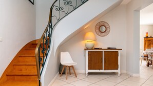 A staircase with wooden steps and a metal railing, a white chair, a wooden cabinet with a lamp, and a dining area with chairs and a table.