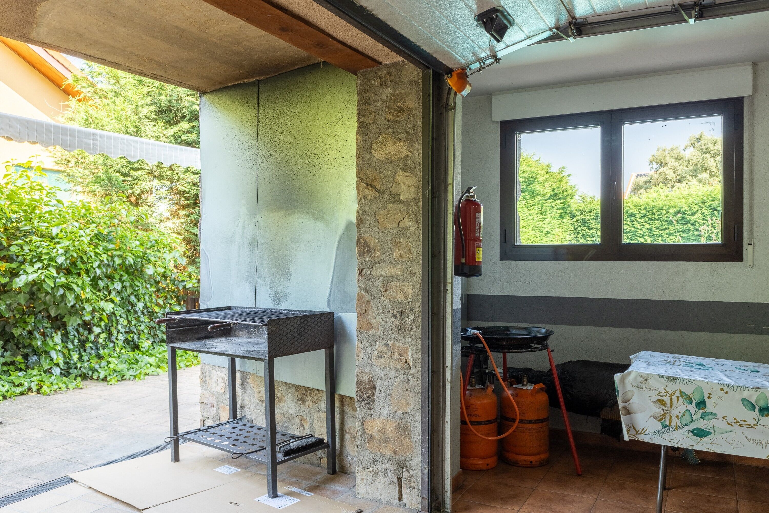 A patio area with a grill, a table with a tablecloth, a fire extinguisher, and a window with a view of greenery.