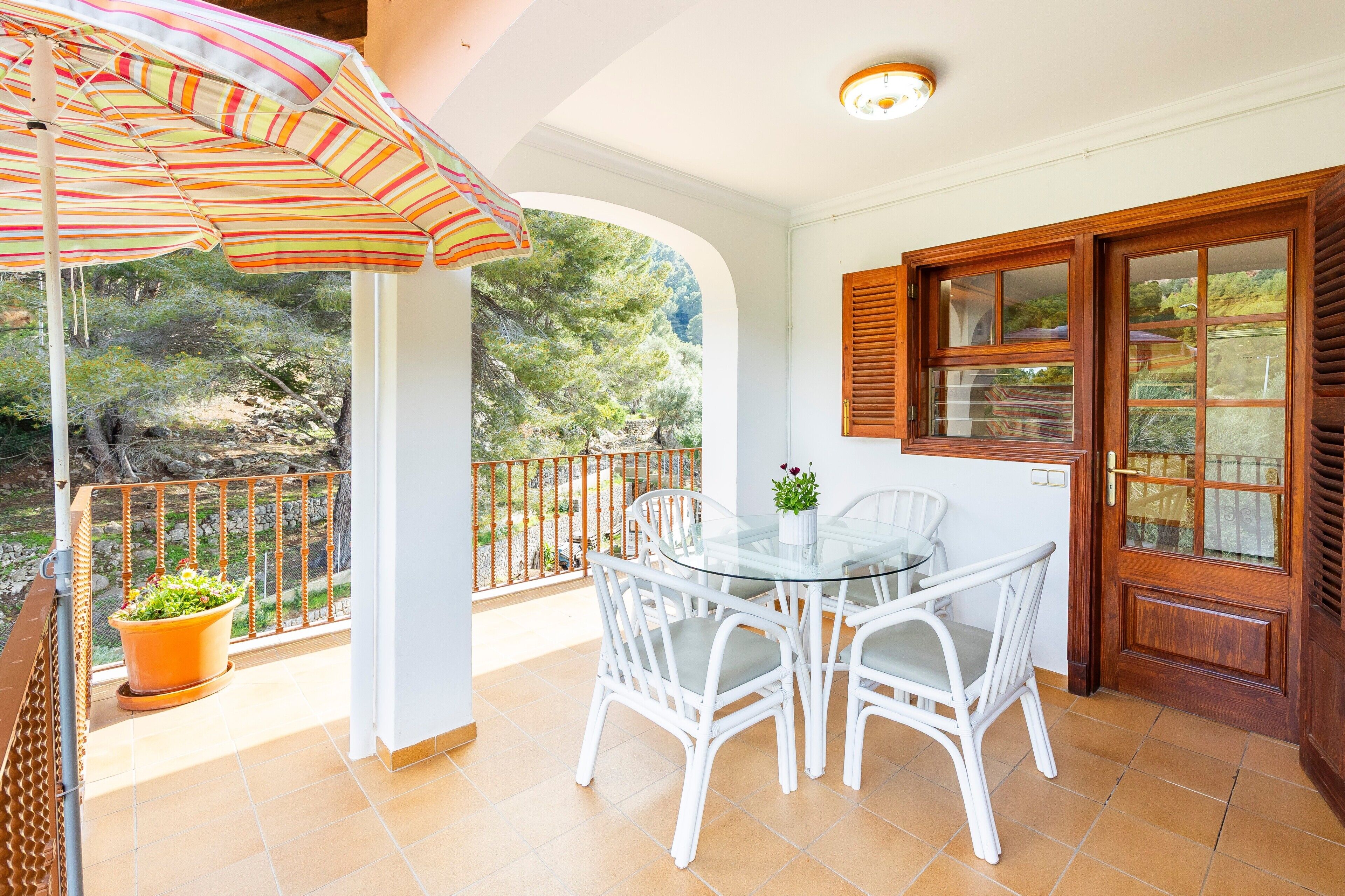 A balcony with a glass table and chairs, a potted plant, and a striped umbrella.