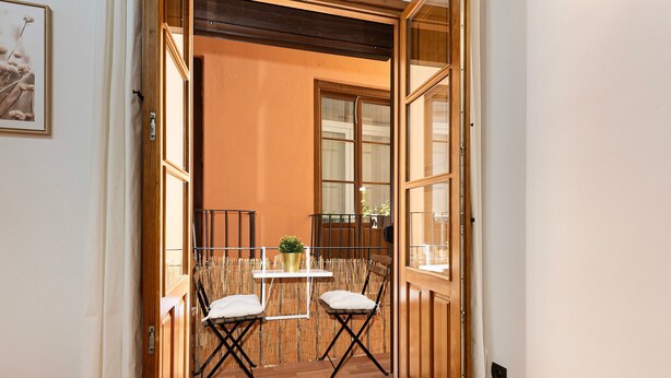 Porta aperta che dà su un balcone con due sedie, un tavolino, una pianta in vaso e vista sull'esterno.