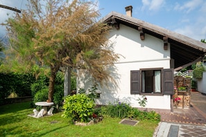 A white building with a brown roof, a small garden with a tree and plants, and a stone bench.