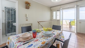 Zona pranzo con tavolo apparecchiato per quattro persone, cabina doccia in vetro e vista sul mare attraverso la porta aperta.