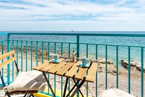 A wooden table with two cups and a coffee pot, a bench with a cushion, and a turquoise railing overlooking the sea.