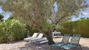 A patio with stone flooring, two lounge chairs, a table, and a tree providing shade.