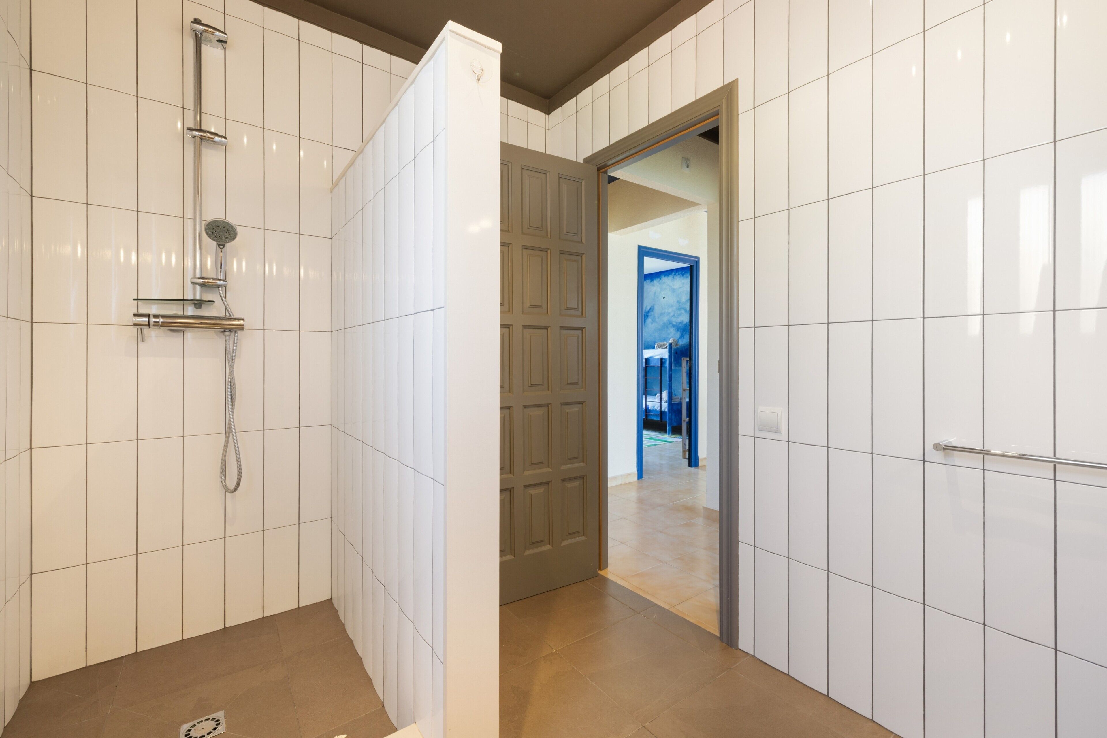 Un baño con paredes de azulejos blancos, una ducha con cabezal de ducha manual y una puerta que da a otra habitación.