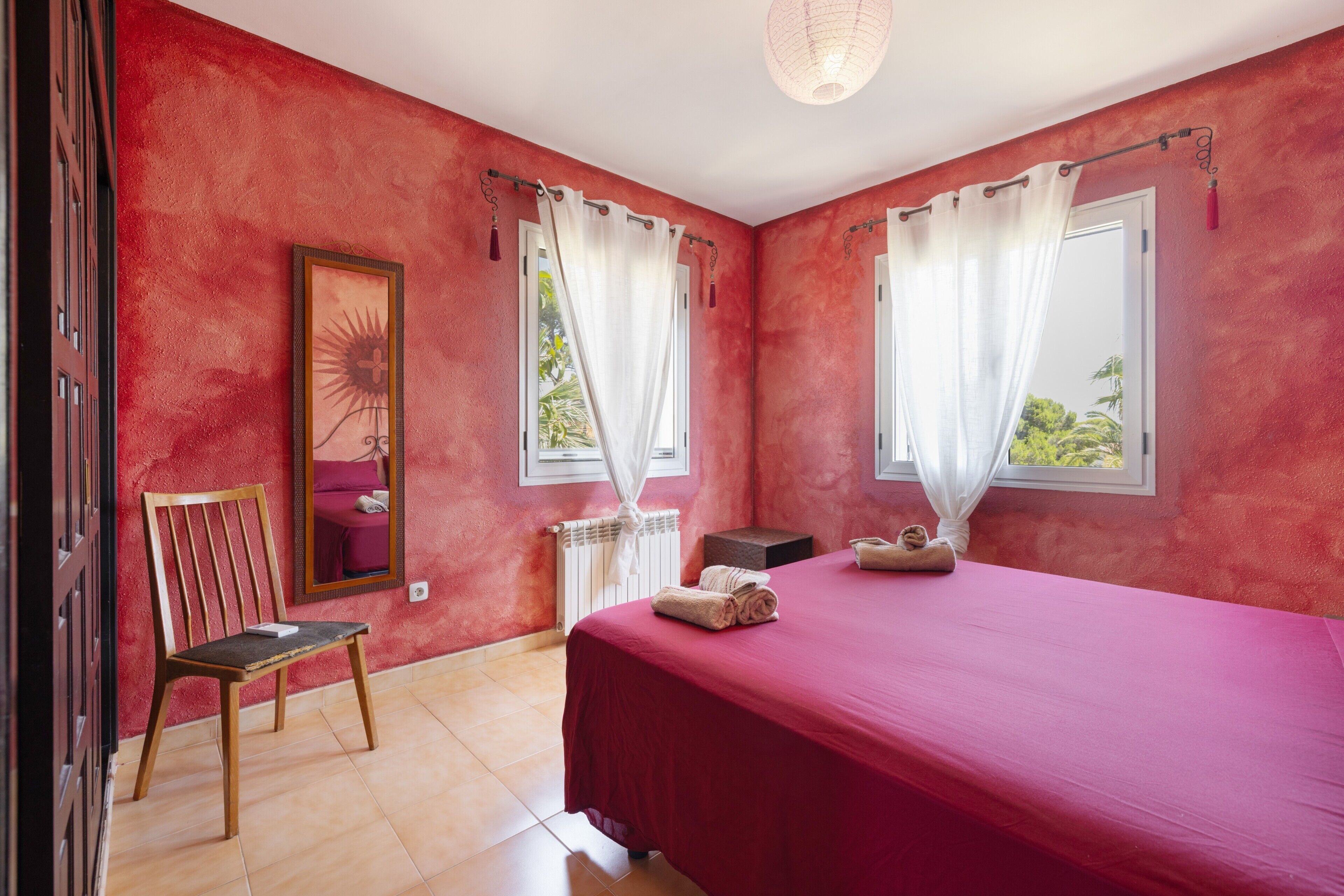 Une chambre à coucher avec un lit, deux fenêtres avec des rideaux, une chaise, un radiateur et un tableau encadré au mur.