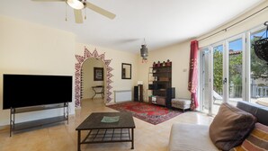 A living room with a flat-screen TV, a coffee table, a sofa, a ceiling fan, a patterned archway, a bookshelf, and a view through large windows.