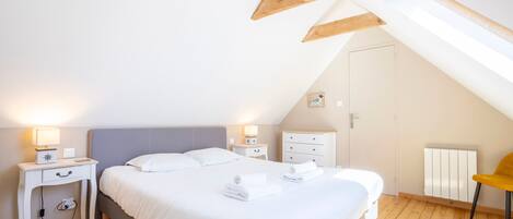 A bedroom with a sloped ceiling, a bed with white bedding, two bedside tables with lamps, a chair, and a radiator.