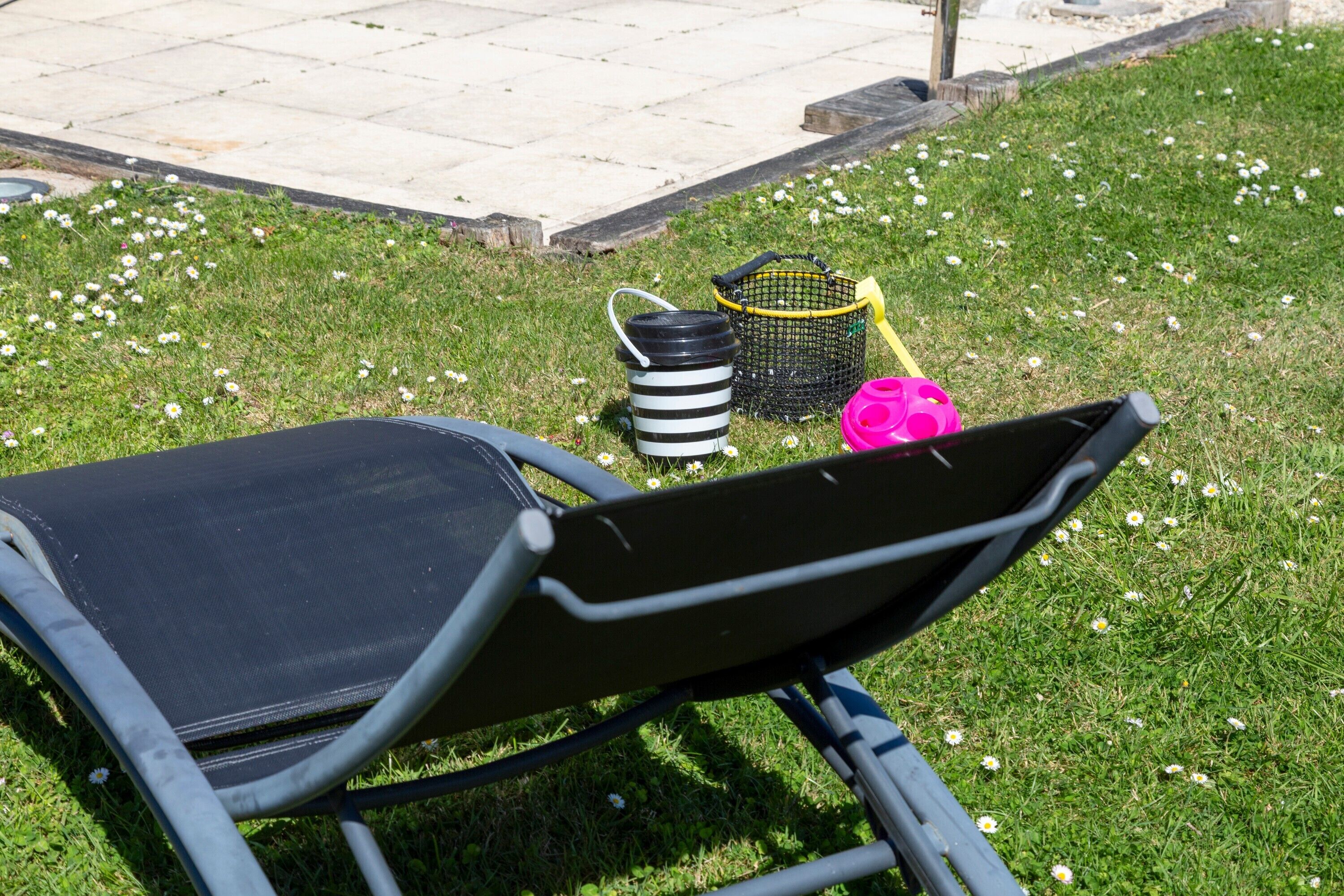 Un jardín con una silla de jardín, un cubo a rayas negras y blancas, una cesta amarilla y un objeto rosa.