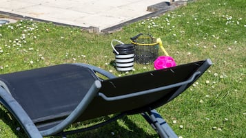 Un jardín con una silla de jardín, un cubo a rayas negras y blancas, una cesta amarilla y un objeto rosa.