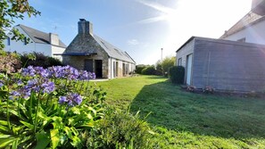 A stone building with a sloped roof, a grassy yard, and purple flowers in the foreground.
